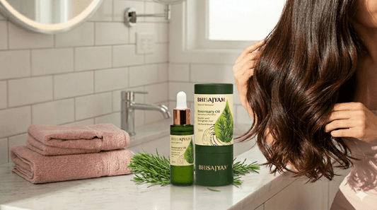 A woman with shiny brown hair stands behind a marble counter featuring Bhisajyam Rosemary Oil and fresh rosemary sprigs in a bright bathroom.