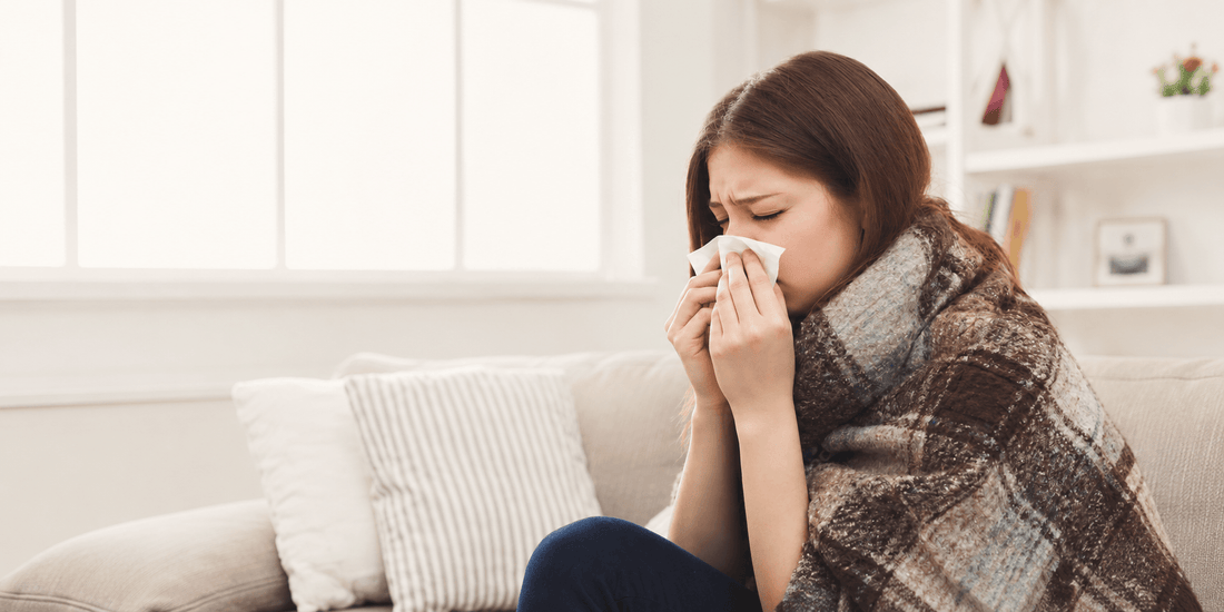Woman wrapped in a blanket sneezing into a tissue while sitting on a sofa, illustrating seasonal cold and flu symptoms during changing weather.